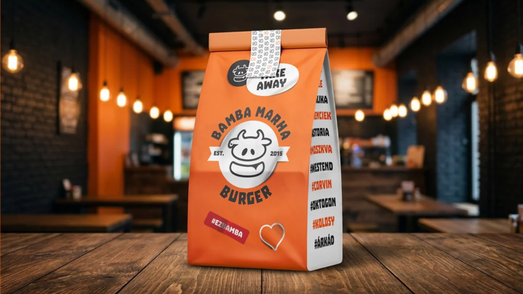 Bamba Marha arculatos elviteles papírzacskó fa asztalon, étterembelső előtt: narancssárga elülső oldalon a mosolygós tehén logó, a Bamba Marha Burger felirat, az EST. 2015 jelzés és a piros #EZBAMBA matrica, tetején TAKE AWAY matricával, oldalán a helyszínek neveivel. A burgerbrand 10 éves rebrandjének csomagolási megjelenése, mockupon.
