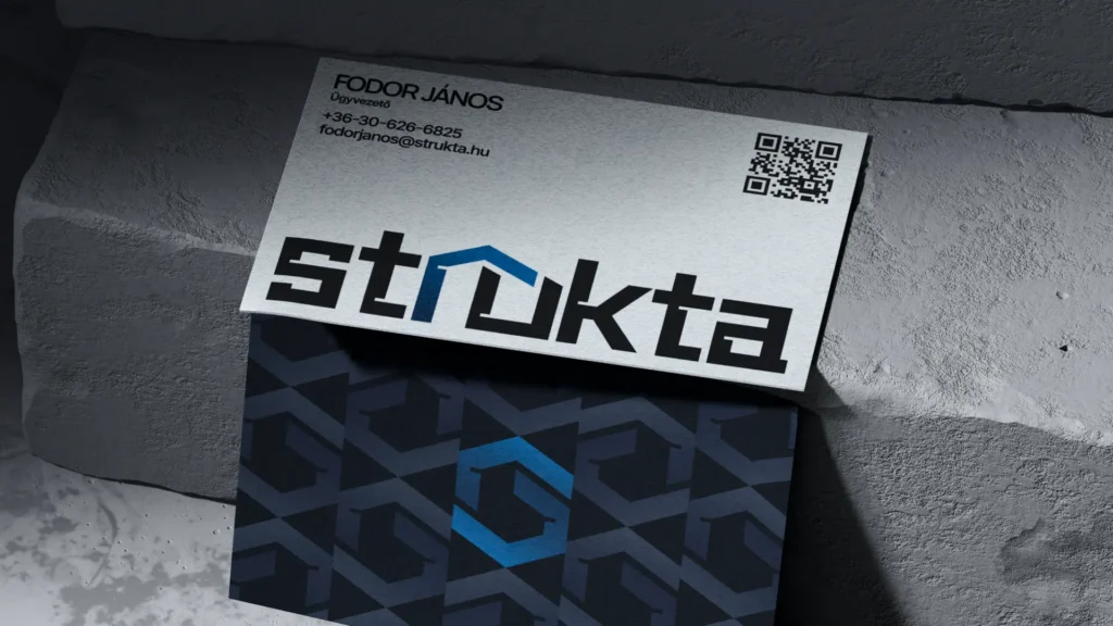 Strukta névjegykártya mockup betonfelületen, mindkét oldalával: az elülső világos oldalon a munkatárs adatai, QR-kód és a nagy STRUKTA logó a kék ék elemmel, a hátsó sötétkék oldalon a márka geometrikus mintázata és a kék hexagon ikon. A generálkivitelező cég arculatának nyomtatott megjelenése.