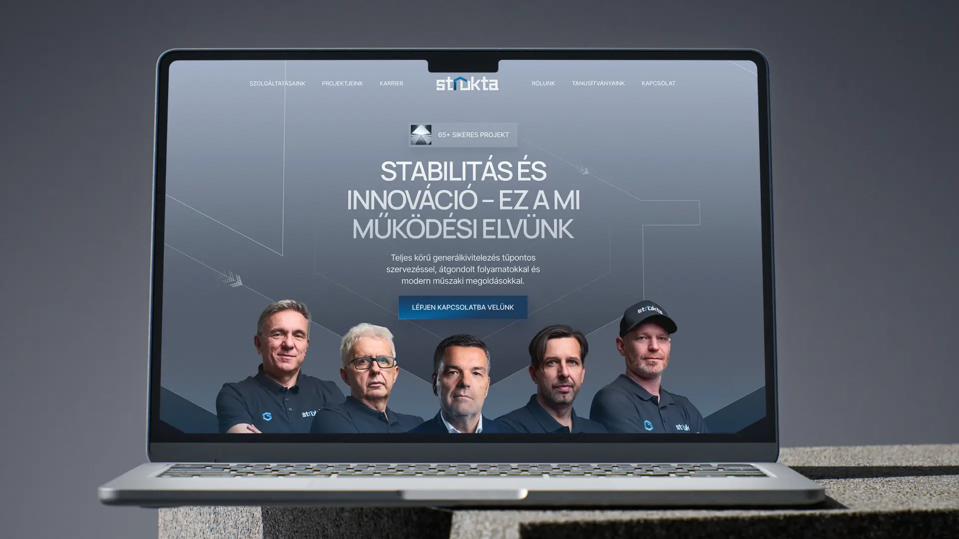 Strukta weboldal-megjelenés mockup egy laptop képernyőjén: a hero szekcióban a márka logója, a „Stabilitás és innováció" főcím, a generálkivitelezést bemutató szöveg és kapcsolatfelvételi gomb, lent a cég munkatársai márkás Strukta-pólóban. A generálkivitelező cég arculatának webes megjelenését bemutató mockup.