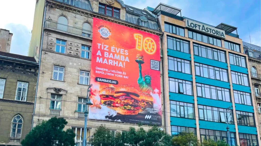 A Bamba Marha 10 éves kampányának óriásplakátja egy belvárosi bérház homlokzatán, az Astoriánál: narancssárga alapon a körlogó, a TÍZ ÉVES A BAMBA MARHA felirat, világító neon 10-es szám, az ünneplő New York-i nyereményjáték üzenete, QR-kód és étvágygerjesztő burgerek. A burgerbrand jubileumi kampányának nagyméretű out-of-home megjelenése.