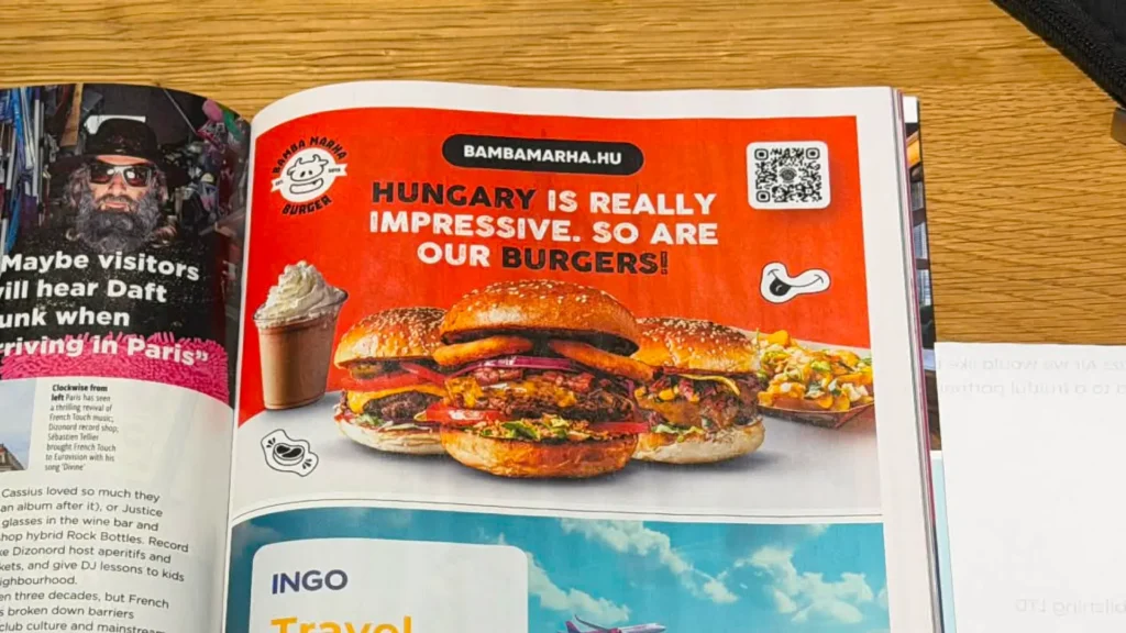 A Bamba Marha angol nyelvű magazinhirdetése a Wizz Air fedélzeti magazinjában: narancssárga alapon a körlogó, a BAMBAMARHA.HU webcím, QR-kód, a Hungary is really impressive főcím, valamint étvágygerjesztő burgerek és milkshake fotója. A burgerbrand 10 éves kampányának nemzetközi közönséget célzó nyomtatott megjelenése.